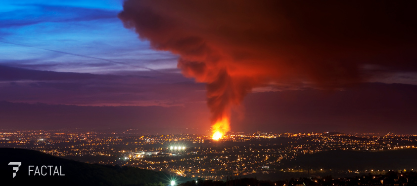 A city at dusk with building lights twinkling across hills. In the center, a massive fire rages, dark smoke billowing into the sky. The Factal logo is superimposed at bottom left.