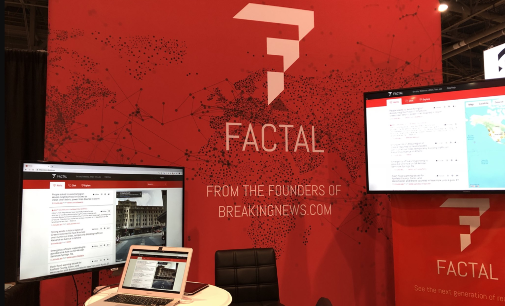 Factal's first booth at GSX. Three screens of varying sizes with Factal's web platform pulled up on each. At back, a red step-and-repeat with the Factal logo and tagline "From the founders of Breakingnews.com" over a map of the world where cities are represented by dots and connected by black lines.