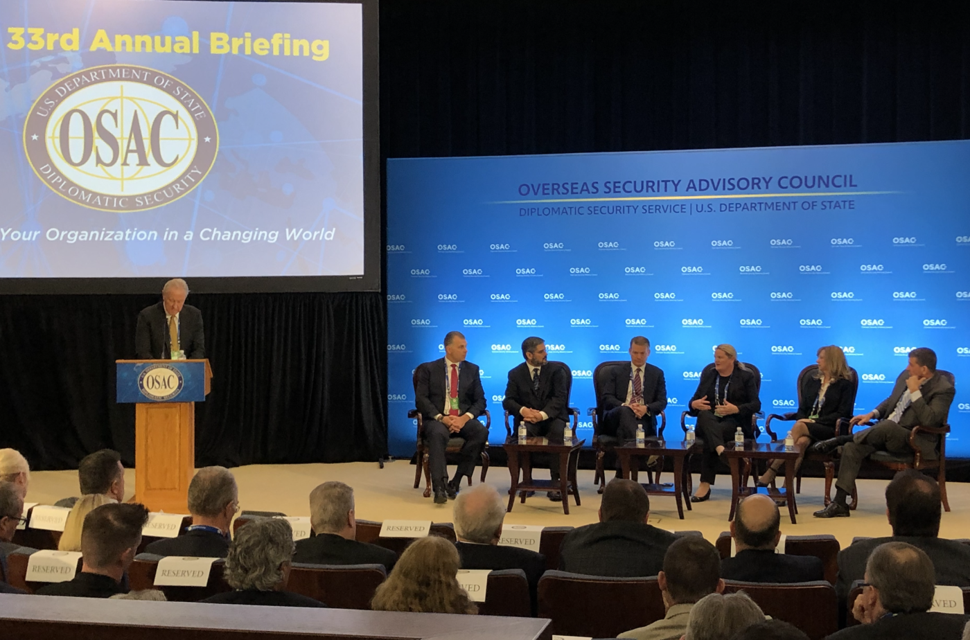 A panel at OSAC. Six people in suits talk animatedly on a stage with a backdrop that reads "Overseas Security Advisory Council." A moderator stands at a podium to the left under a screen with "33rd Annual Briefing - OSAC - Your organization in a changing world" projected onto it.