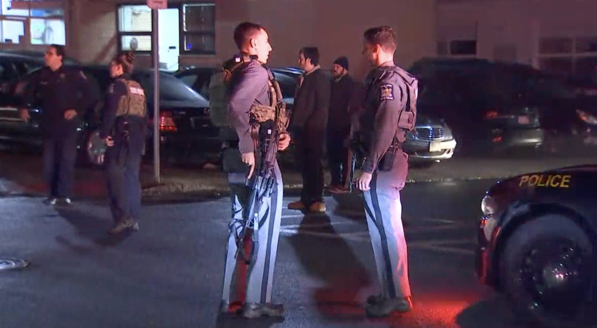 Law enforcement personnel and civilians stand and talk in a parking lot after dark.