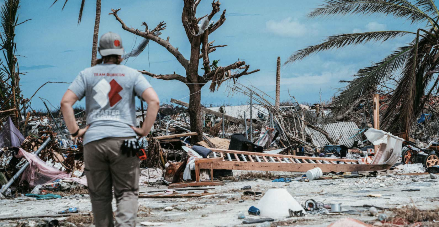 A member of NGO Team Rubicon stands arms akimbo looking at rubble left behind in the wake of a storm on a beach.