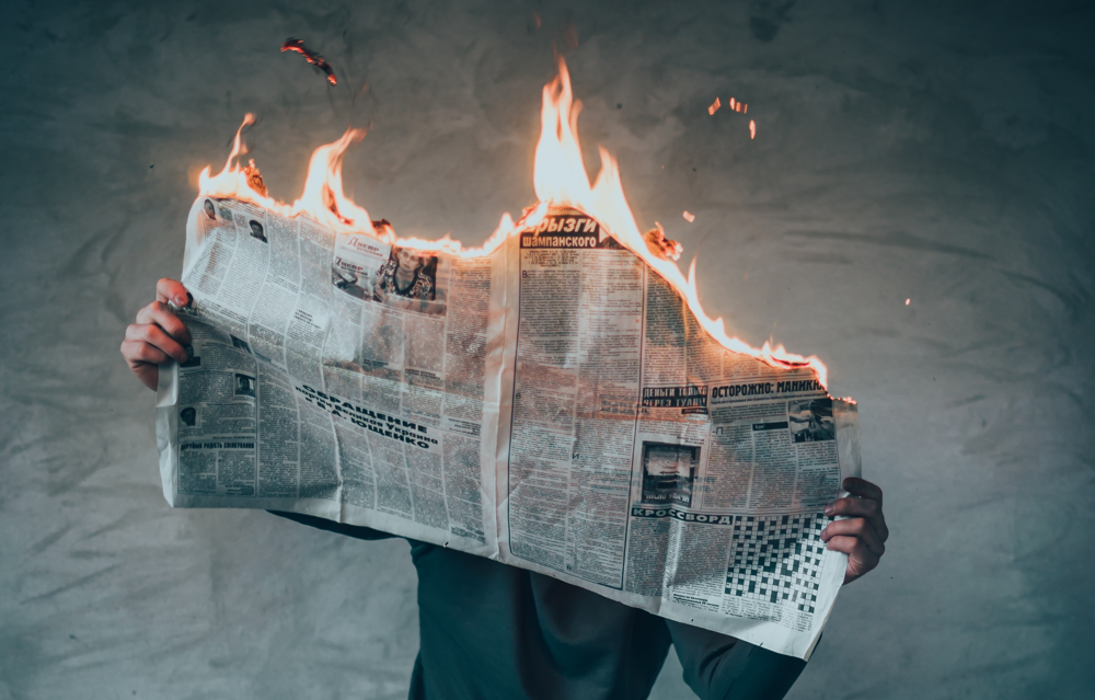 Image of a person holding a Russian newspaper open in from of their face. The paper is burning from the top down.