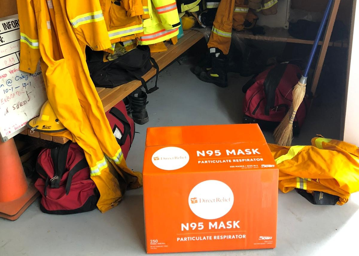 A large orange box labeled "Direct Relief: N95 Mask, Particulate Respirator, 250 count" sits in a ready room next to first responders' uniforms.