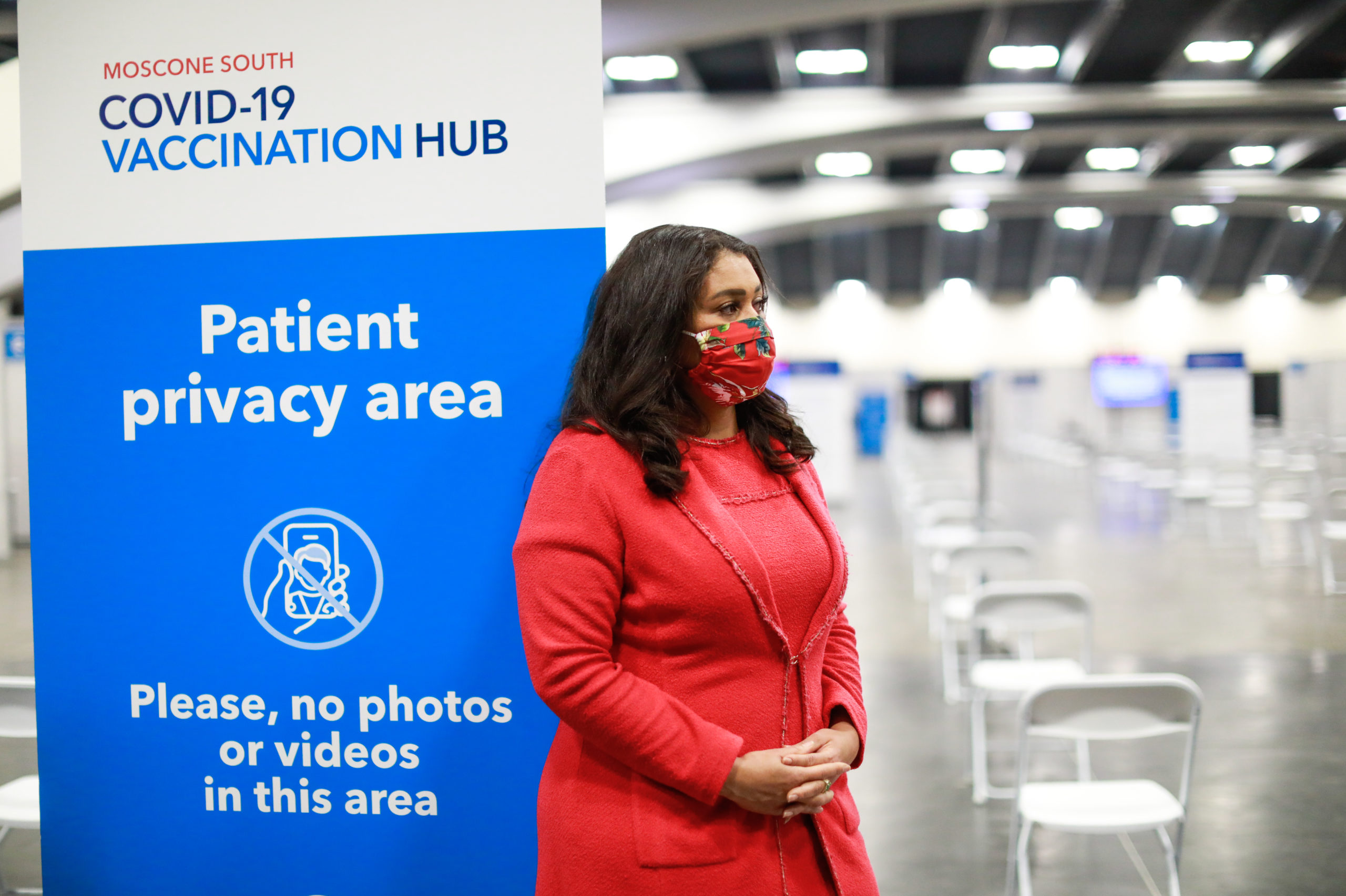 A single individual wears a cloth mask in an empty room. She stands next to a sign that says Patient privacy area, no photo or videos