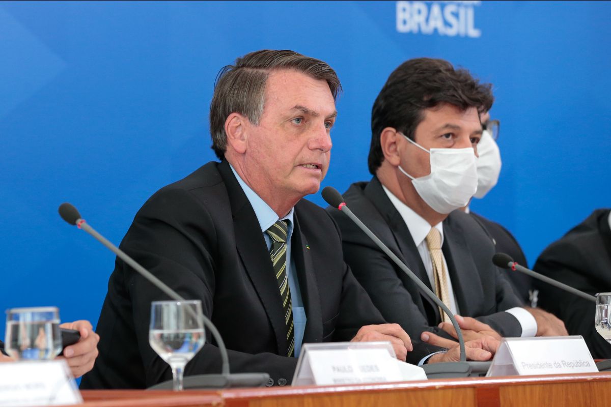 Jair Bolsonaro speaks into a microphone at a dais, flanked by men in suits wearing masks - Bolsonaro is not wearing one.