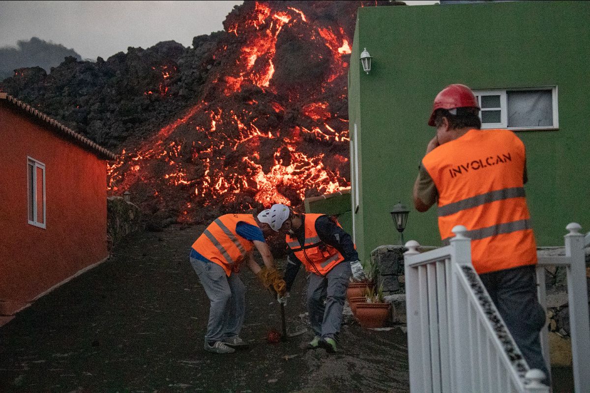 Workers wearing safety gear and bright ornage vests stand between two one-story residences -- while molten, glowing lava looms in close proximity to them.