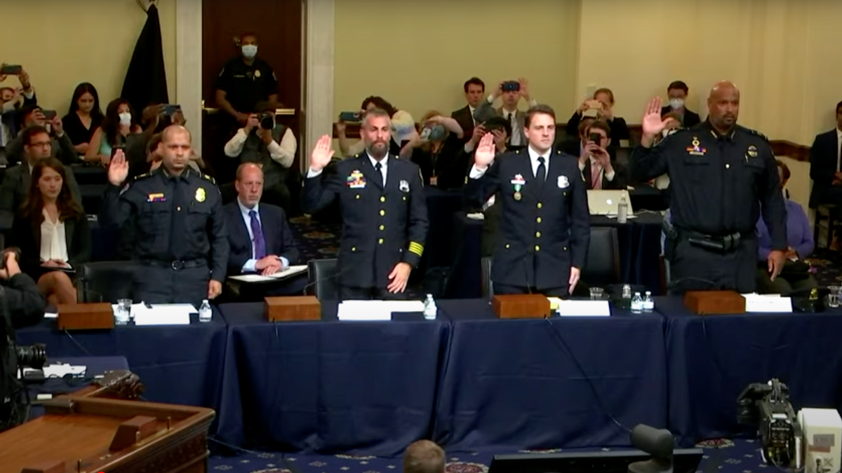 Four uniformed officers stand and raise their hands behind desks covered in blue tablecloths in a crowded room.