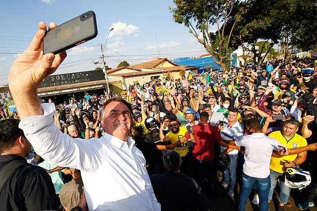Jair Bolsonaro takes a selfie in front of a throng of enthusiastic supporters, a few wearing masks, on a sunny day.