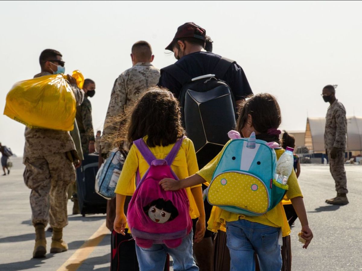 Adults in military garb walk along a tarmac, followed by two small children wearing colorful backpacks.