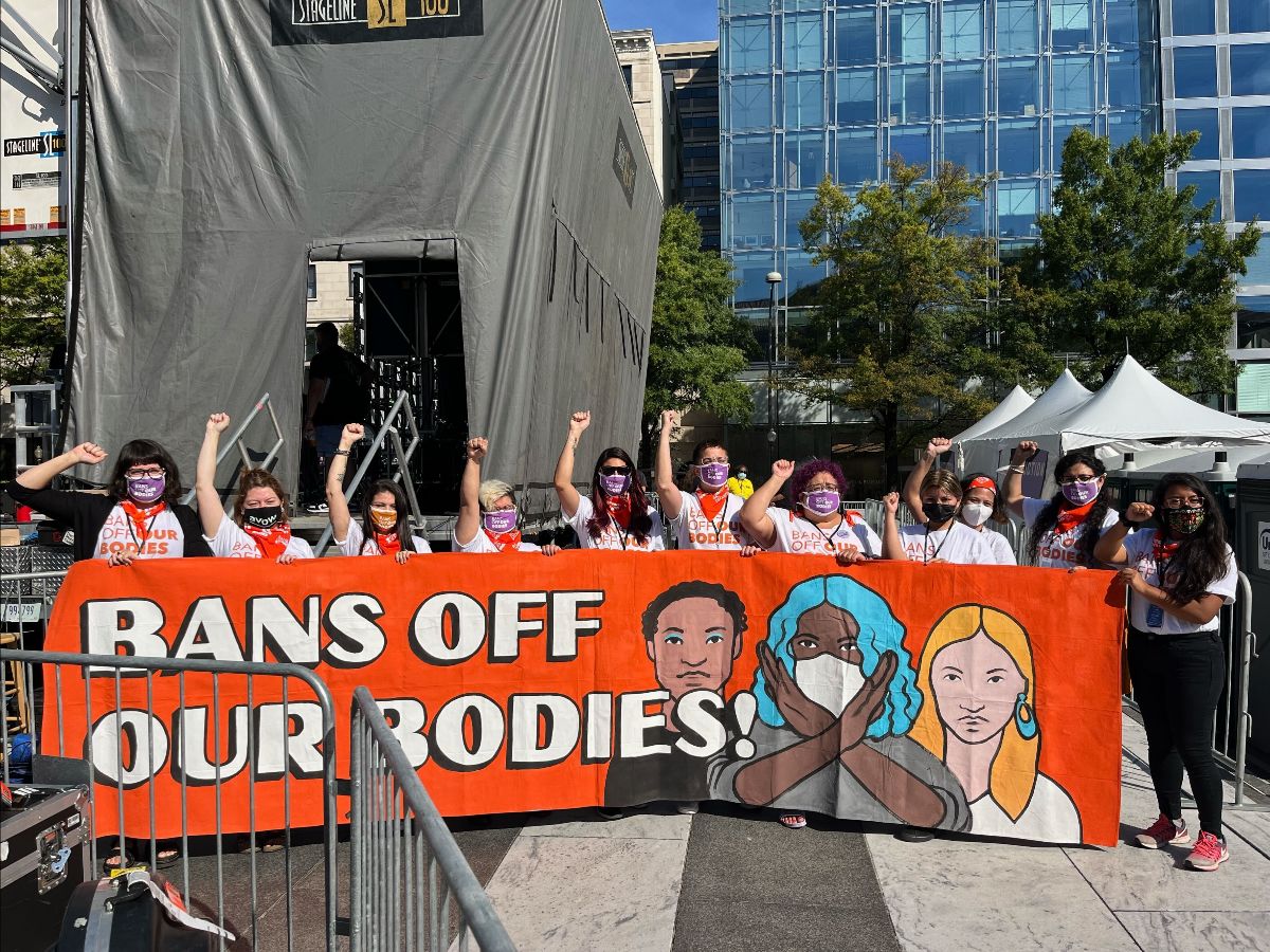 Eleven masked protesters raise their fists behind a sign that reads "BANS OFF OUR BODIES!"