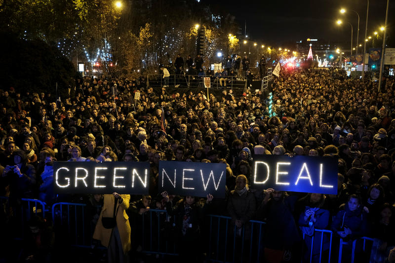 Masses of people stad shoulder to shoulder at night on a city street. An illuminated sign in three pieces, held by three people, reads "GREEN NEW DEAL".