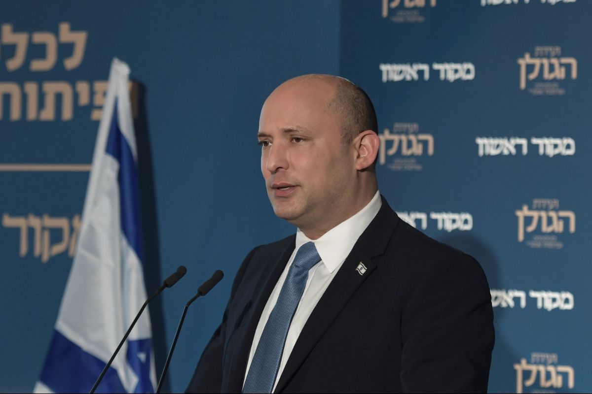 Naftali Bennett stands in front of a backing with Hebrew words