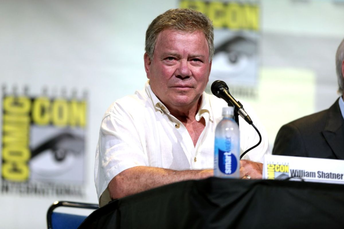 William Shatner, an older white man, sits at a table, looking at the camera, wearing a loose-fitting white shirt