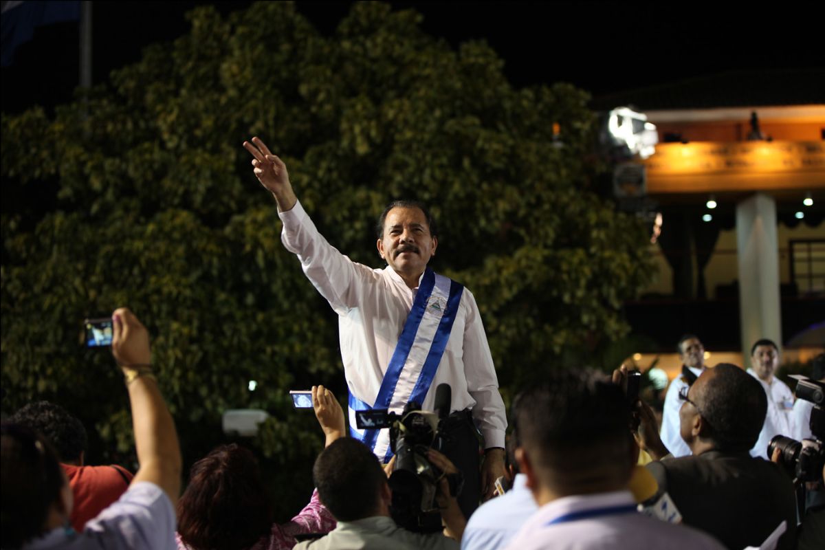 Daniel Ortega, a mustached man wearing a white dress shirt and blue-and-white sash, gestures to a crowd.