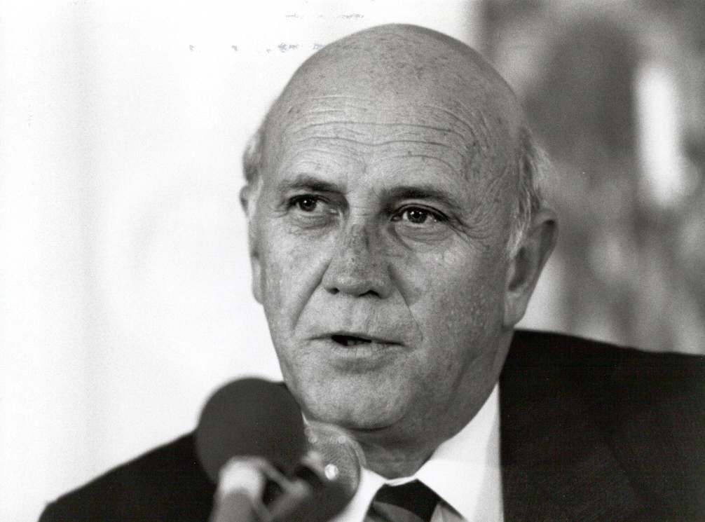 A black-and-white closeup of a mostly bald white man, wearing a suit, speaking into a microphone.