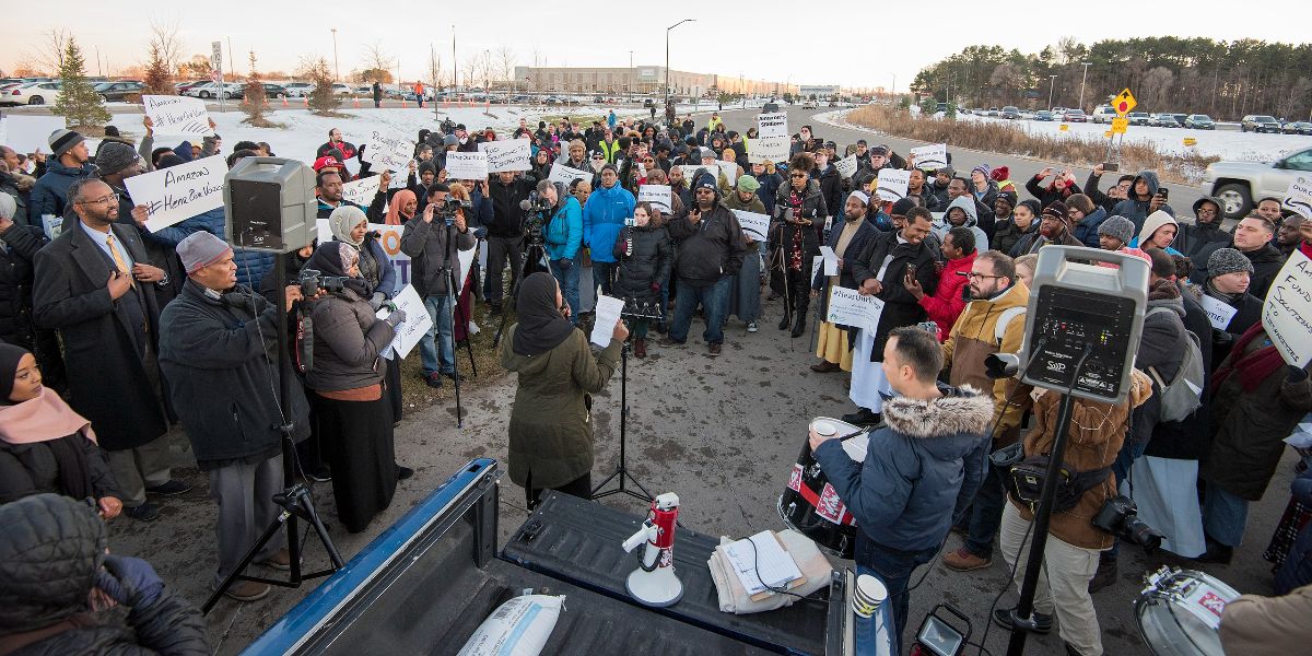 People stand in a circle, some holding signs, as someone holds a sheet of paper and speaks into a microphone. Snow is visible on the ground.
