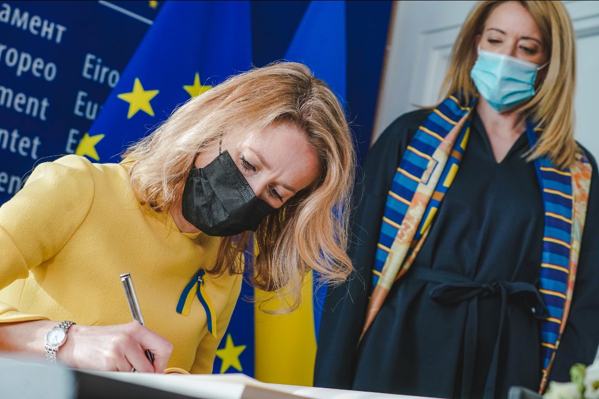 Two members of the EU parliament are featured. The one on the left is in yellow, signing a document. Behind her is another woman, in a navy blue top with a blue and yellow scarf on. Both are wearing masks. Behind them is an EU flag.