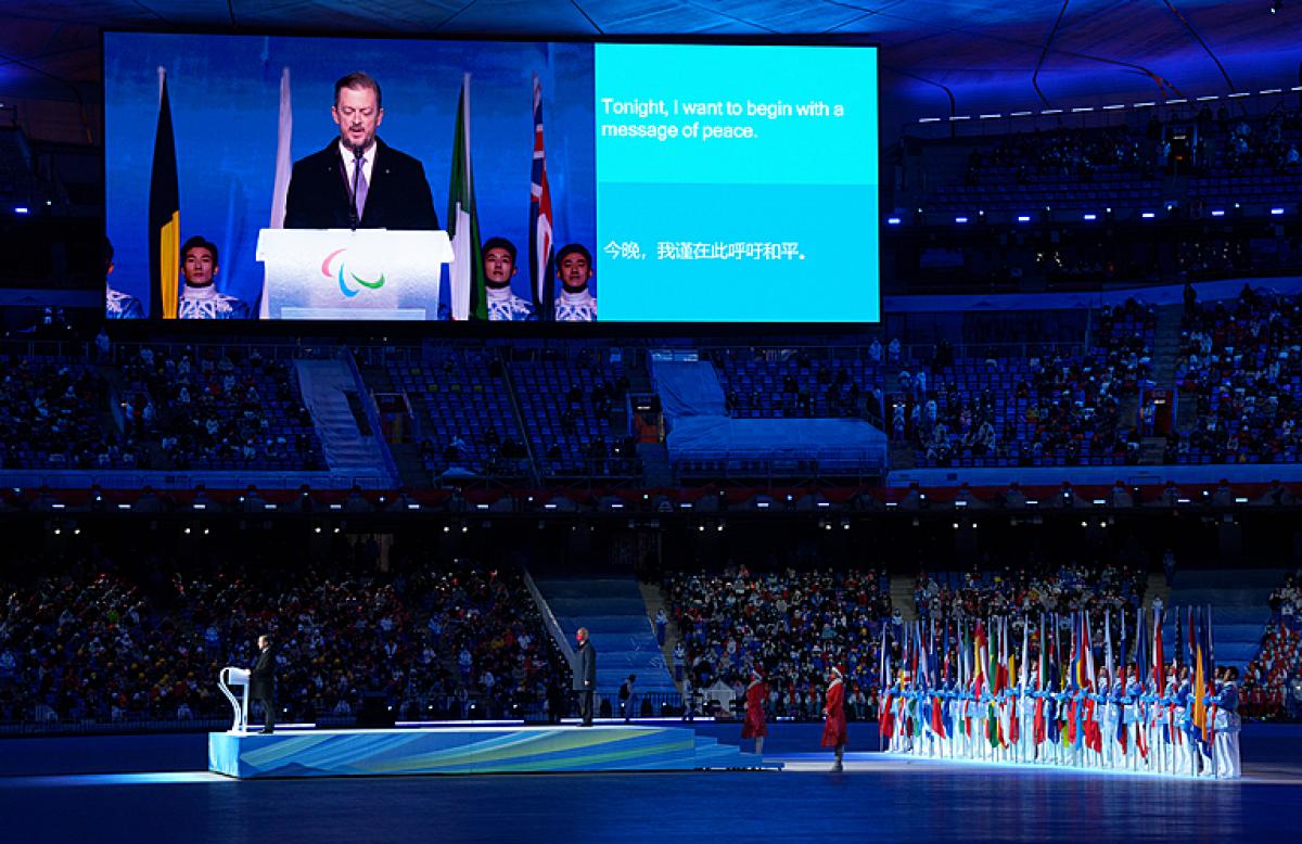 A wide photo of the opening ceremony for the 2022 Paralympics in Beijing. On a dais is the IPC president and one other dignitary. To the right of the image are dozens of people holding flags of the participating nations. The whole scene is in a large stadium with stands full of athletes, coaches, and administrative officials.