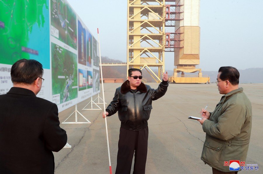 North Korean leader Kim Jong un stands in front of the framework of a rocket launchpad. Besides him are maps and diagrams. He is holding a long stick as if ready to point at specific items.