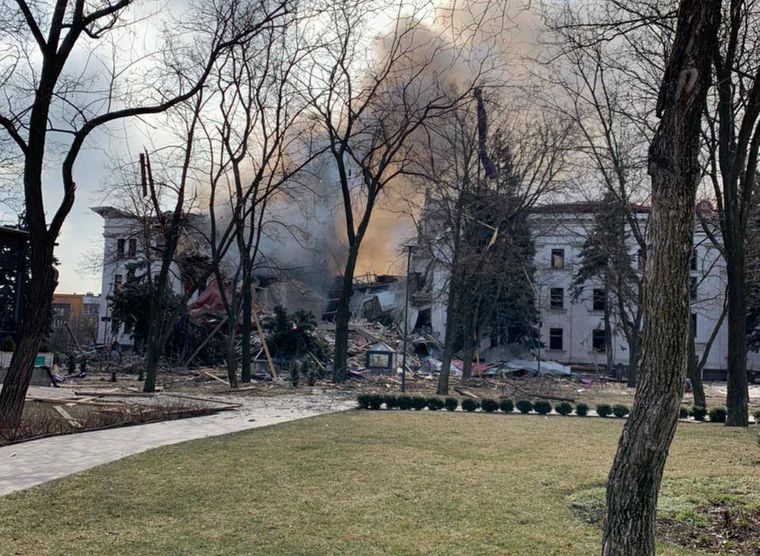 a photo of a theater in Mariupol, Ukraine showing extensive damage from an airstrike. The center of the building is destroyed with the right side having less damage than the left. The foreground shows a lawn. Nearer to the building are trees by the damaged building. The trees are knocked over.