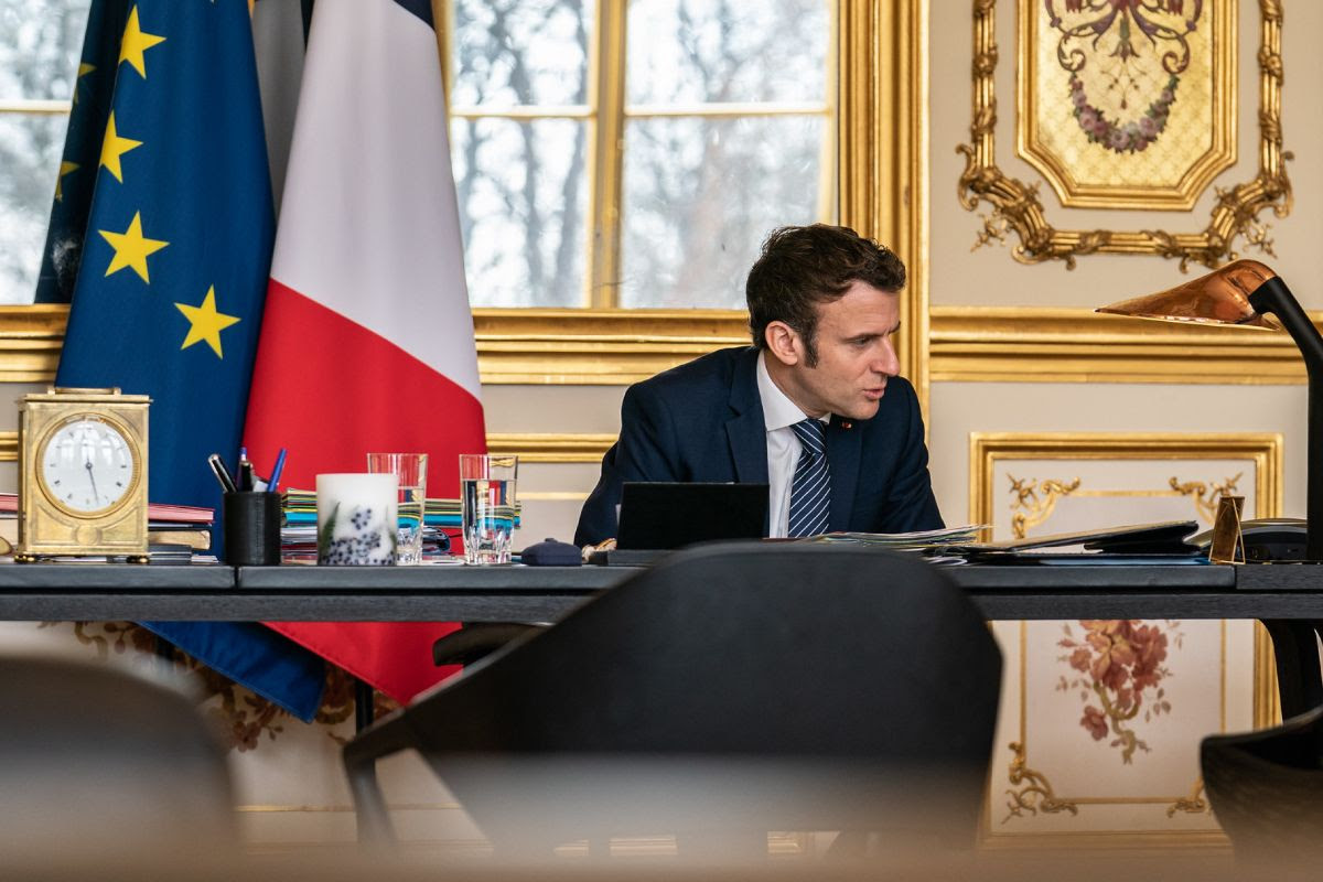 French President Emmanuel Macron speaks by phone at his desk. Behind him are the flags of the European Union and France.