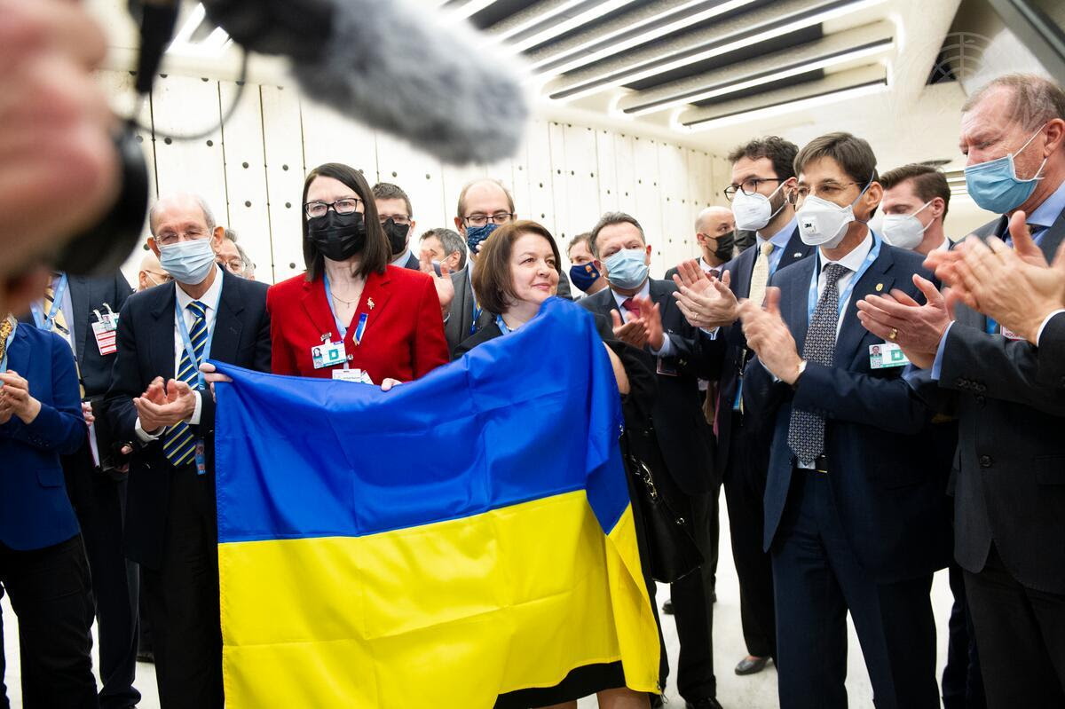 A couple dozen diplomats in suits wearing N95 or surgical masks stand applauding Yevheniia Filipenko, Ukraine's permanent representative to the United Nations as she stands behind the flag of Ukraine.
