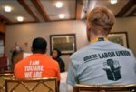 Two people sit in a room listening to a speaker. Both seated individuals are wearing pro-union t-shirts, one in orange and the other in grey.