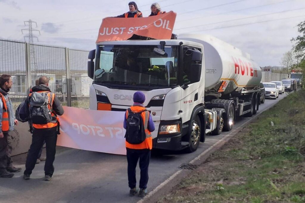Four protesters stand in front of an oil delivery truck as it drives down the road. Two of them face the truck holding a sign that the driver can read. Two additional protesters are on top of the cab of the track. They also hold a sign, this time it is facing the viewer. The sign says "Just Stop Oil"