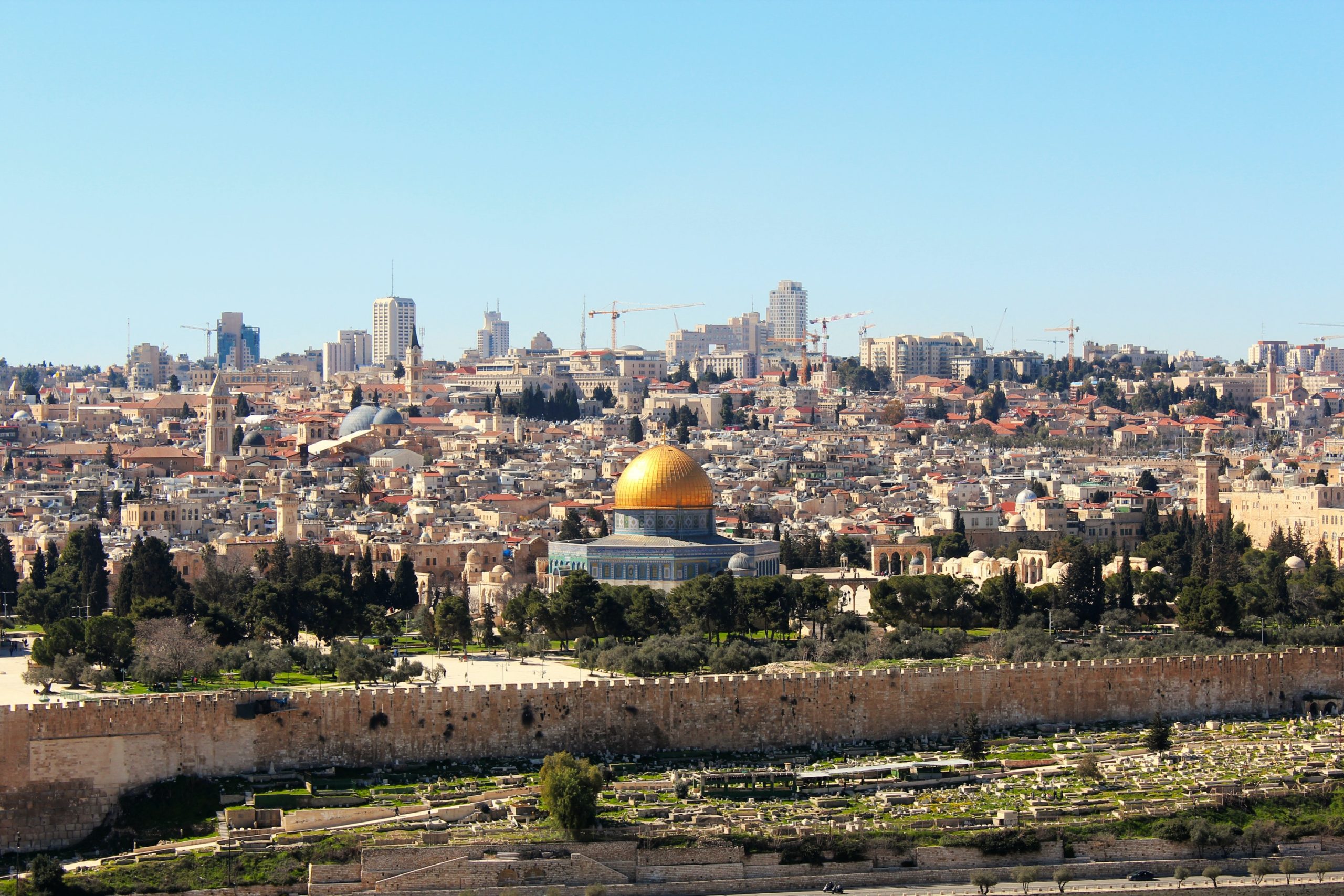 A long distance photo of Jerusalem. In the foreground is a cemetery. In the midground is the Dome of the Rock and the Old City. Beyond those structures are modern buildings and barely visible, due to distance, construction cranes.