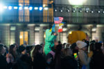 Dozens of people stand in a street all facing towards the same speaker. Mostly in dark warm clothes the central person has a small child in a teal jacket. The child is on the shoulders of that individual and waving flags of Poland, the United States and Ukraine.