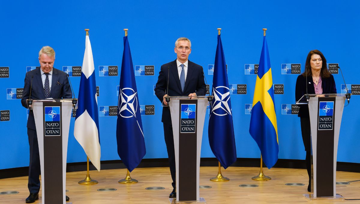 In the center of the photo is the NATO Secretary General. To the left of the photo is Finland's Foreign Minister Pekka Haavisto and to the right is Sweden's Foreign Minister Ann Linde. The three people stand in front of four flags. The flags of the nations are behind their foreign ministers. In the center are two NATO flags.