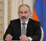 Nikol Pashinyan sits at a desk while wearing a dark suit jacket over a white shirt with a grey tone tie. He is seated in front of the Armenian flag.