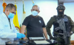 Dairo Antonio Úsuga is in the center of a photo. He, the inprocessing person to the left and the guard to the right, is wearing a medical mask. The guard is armed and in uniform. The inprocessing person is wearing a lab coat.