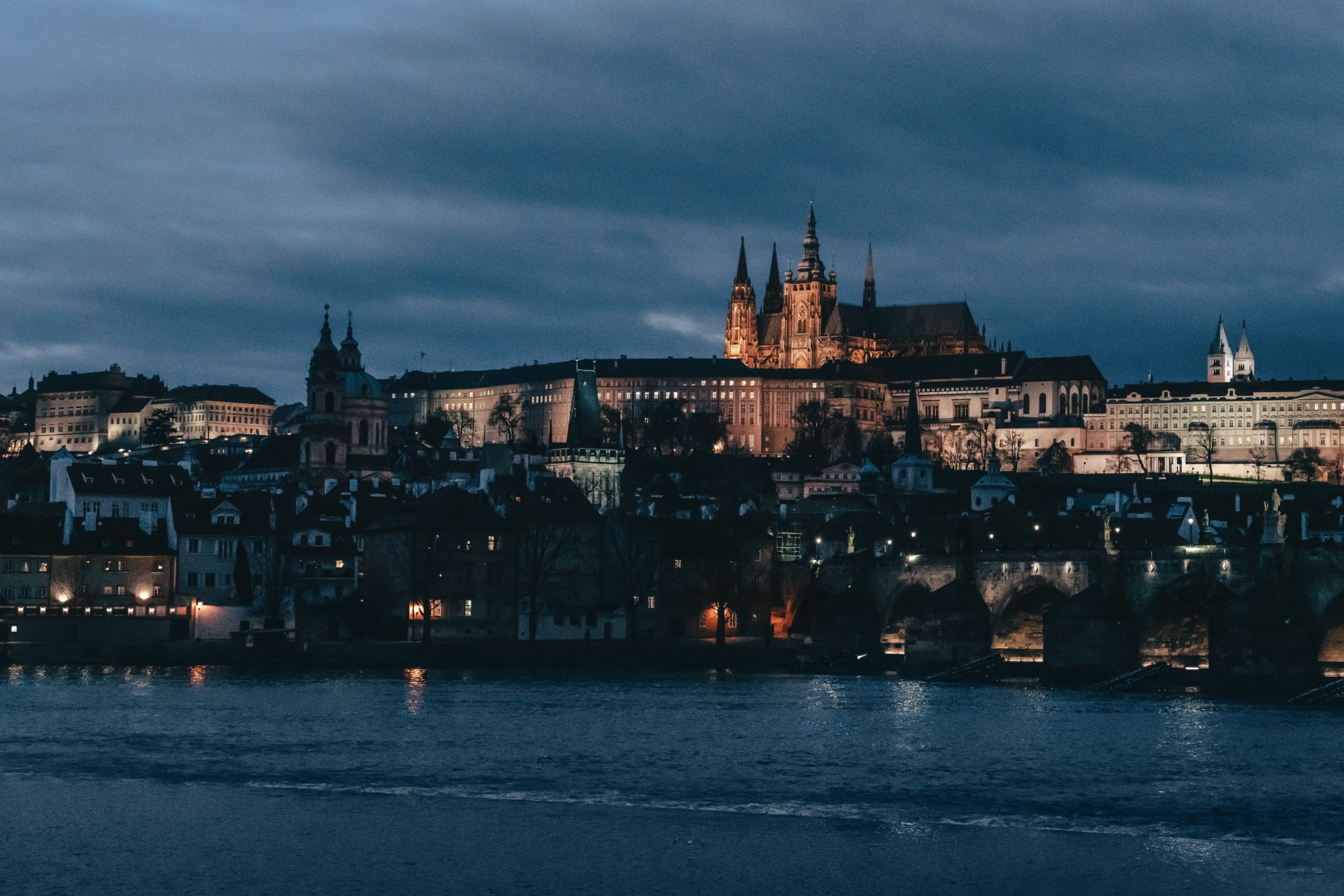 A photo of Prague Castle looking across the river. The photo is taken in the early evening with a deep, dark blue sky and light cloud cover. Most of the buildings have lights on. The castle is lit from the ground.