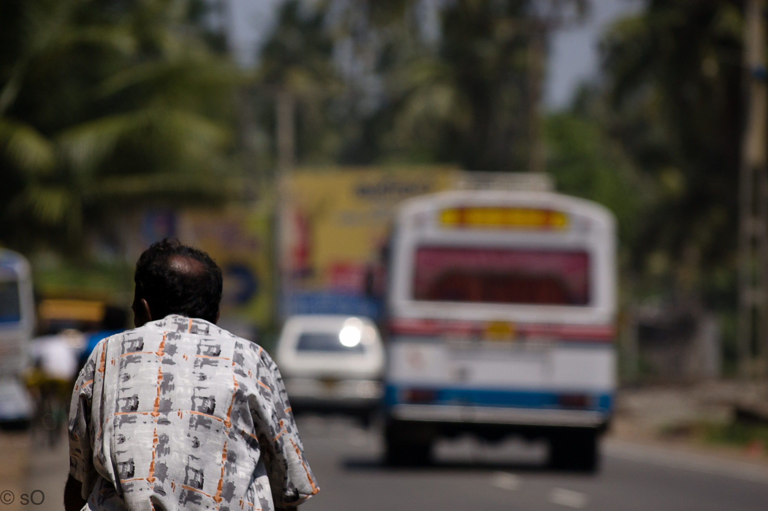 A man, either seated or riding a bicycle, is in focus. The photo is of his back as he looks at a bus on a Colombo, Sri Lanka road.