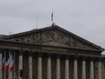 A photo of Palais Bourbon. The seat of the French National Assembly has a front with many columns and a medium peaked roof. There are relief statues under the roof. It is topped by a French flag and some antennas. Beside the grand entry are three more French flags.