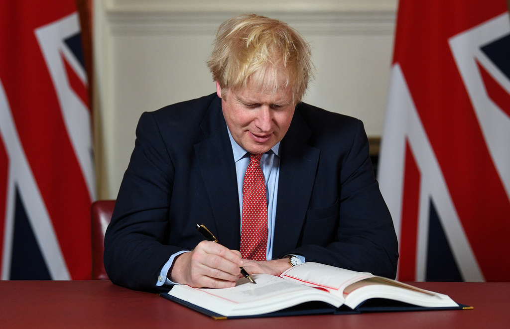Prime Minister Boris Johnson sits at a desk and signs the U.K. withdrawal agreement. Behind him are two United Kingdom flags.