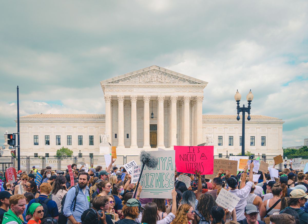 Protesters gather outside the U.S. Supreme Court building. Between them and the building are two sets of fencing and at least one guard with a reflective vest on. The protesters are mostly facing to the right as if walking that direction or focused on a speaker.