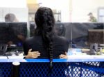A photo of the back of a women's head, her hair in braids. There are small hands of a child on her back as they hug. The two sit on a metal bench in a facility that processes migrants.