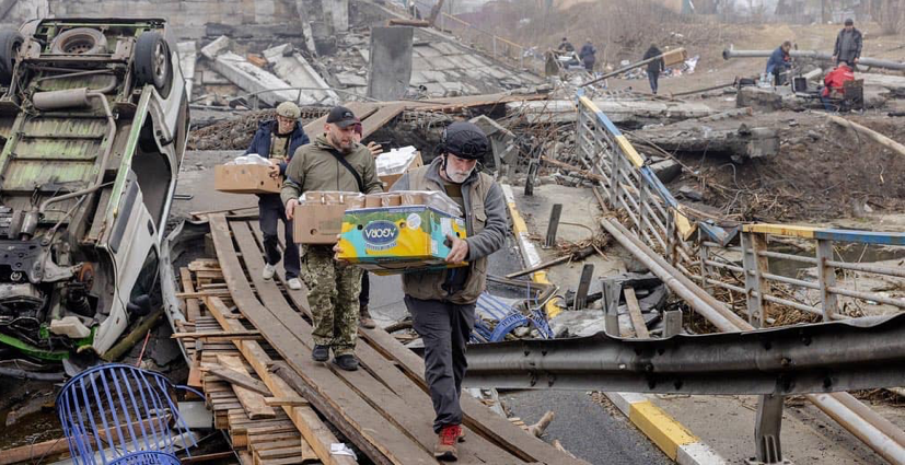 four individuals carry goods across a make shift bridge. There are overturned cars and destroyed buildings to their sides.