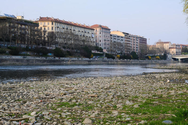 A photo of of a river and residential buildings in a town in northern Italy. In the foreground is a river with very little water, a rocky shore close to the camera's viewpoint. Beyond the river are a collection of 6-8 story buildings.