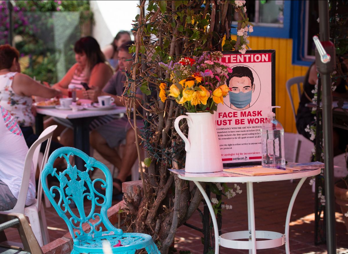 An outdoor restaurant with a sign on a table requesting that people mask. A table in the background shows a couple people without masks.