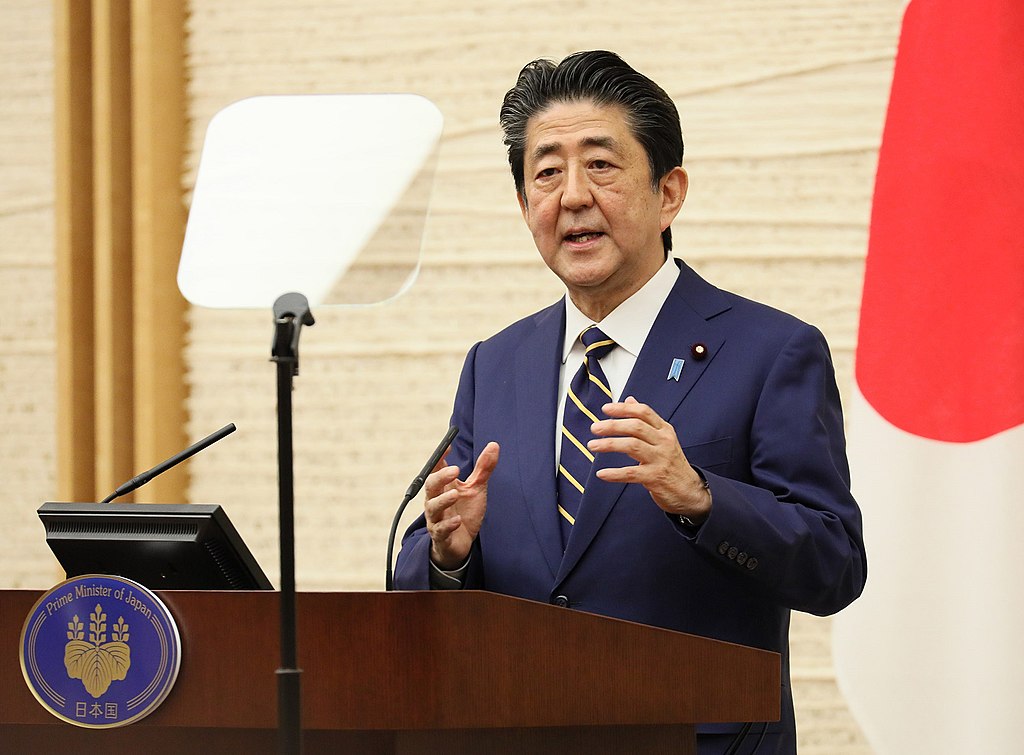 Shinzo Abe, former Primer Minister of Japan, stands at a lectern delivering a speech. He is in a suit and tie.