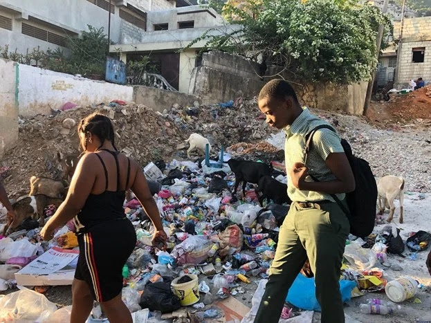Two individuals walk from right to left in front of a pile of trash in Port-au-Prince, Haiti. 