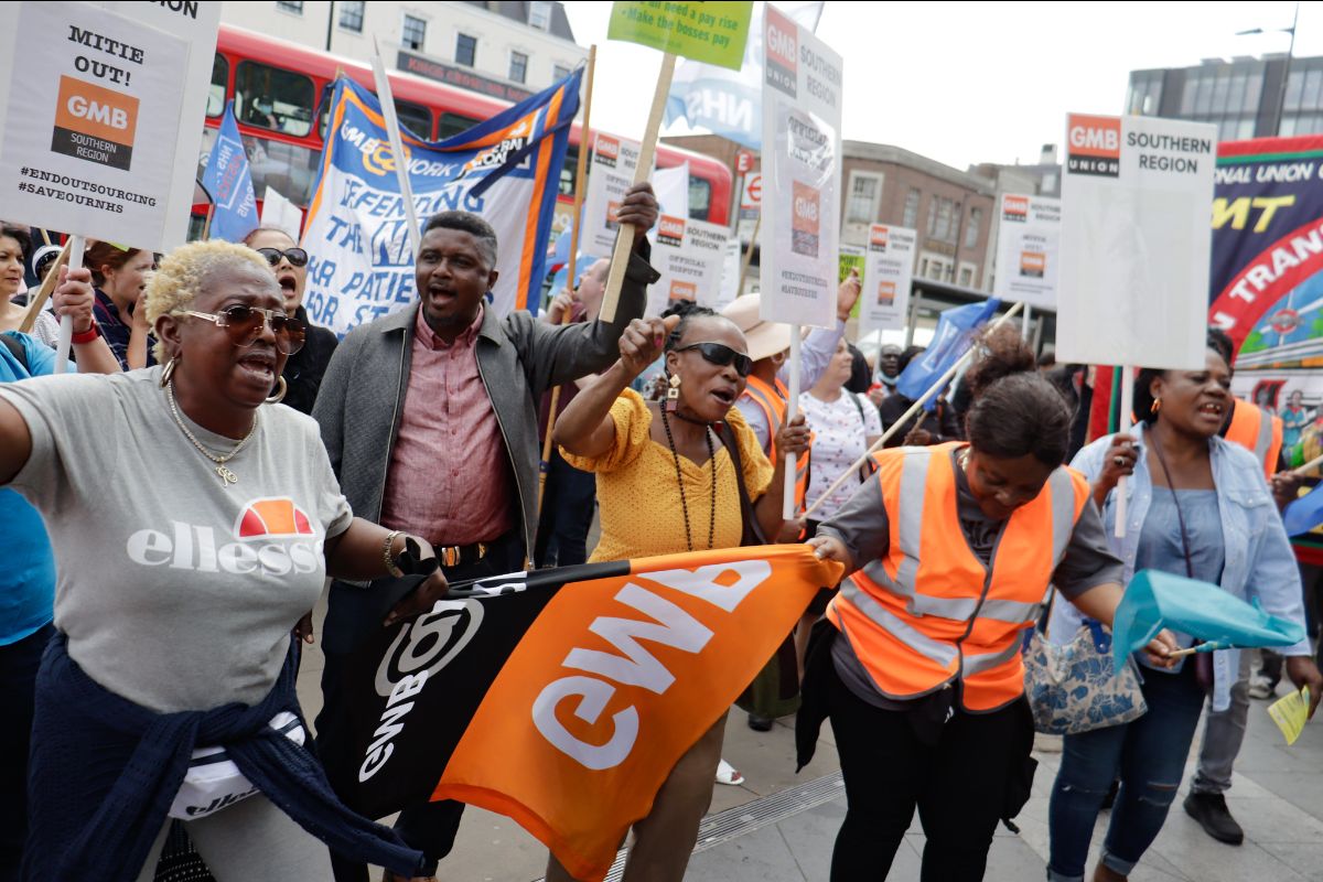 A closeup photo of demonstrators outside of King Cross Station in London. There are five people in the front row, all have banners or signs in hand. Behind them there are dozens and dozens more people. None of the clothing is a uniform; the people only wearing civilian attire. The signs almost all say GMB.