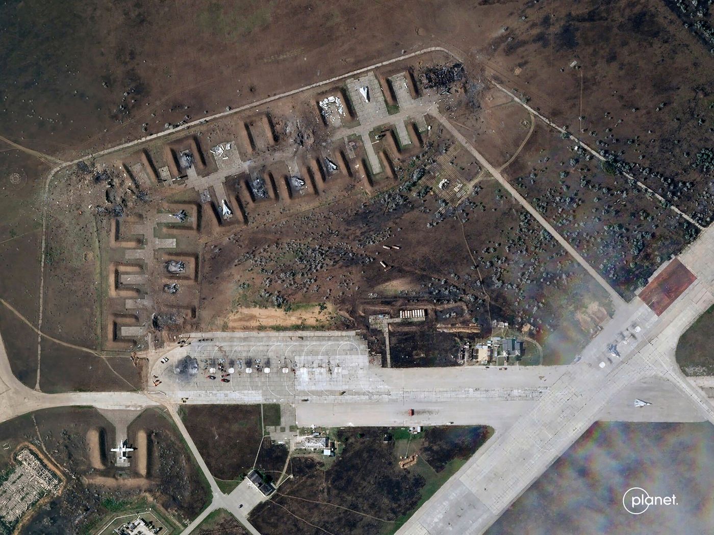 This satellite image of a military air port in Crimea shows significant damage to the main building and some outbuildings and several protective bunkers for fighter jets. The two runways have scattered debris but little damage.