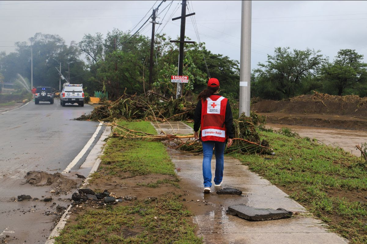 An aid worker with American Red Cross vest on, walks down a rainy sidewalk that is blocked by debris from Hurricane Fiona