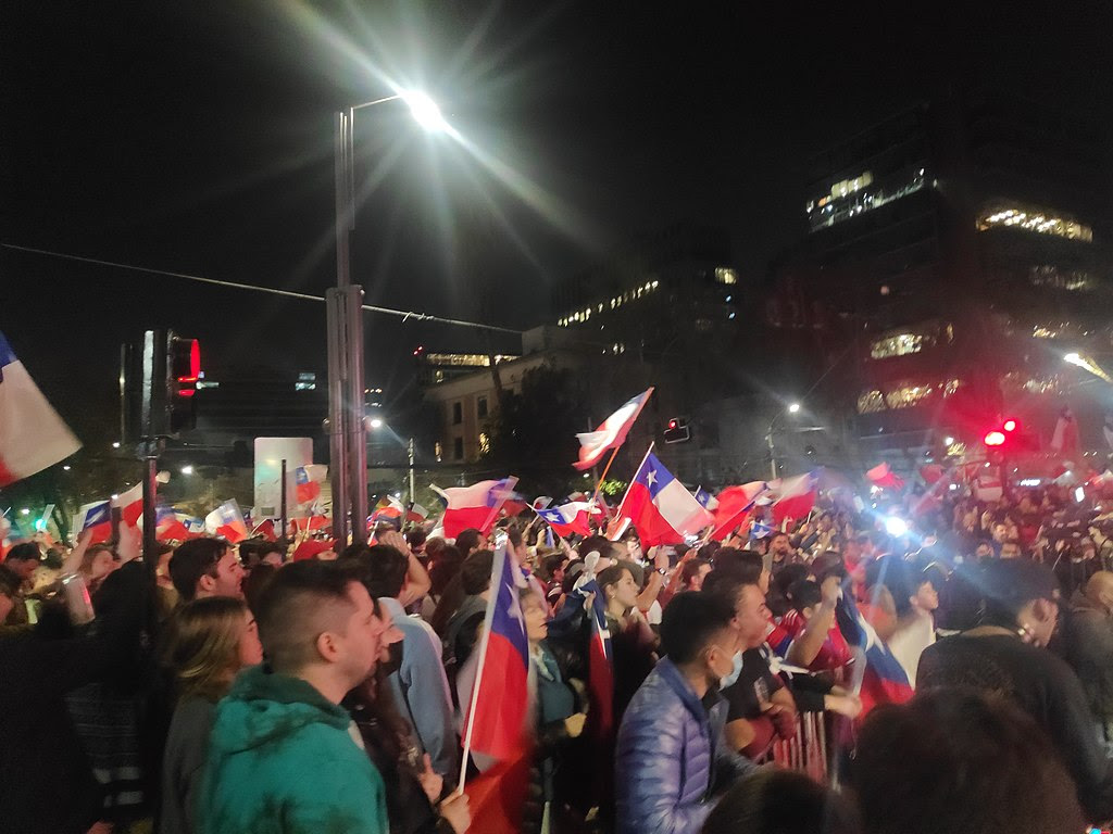 A photograph of a crowded street at night. There are hundreds of demonstrators on a wide street or plaza. A few dozen are flying the flag of Chile.