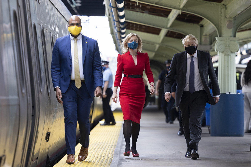 A photo of three people on a train platform. In the middle is Liz Truss, in a red dress. To her right is former UK Prime Minister Boris Johnson in a navy suit. To the left is an unnamed man.