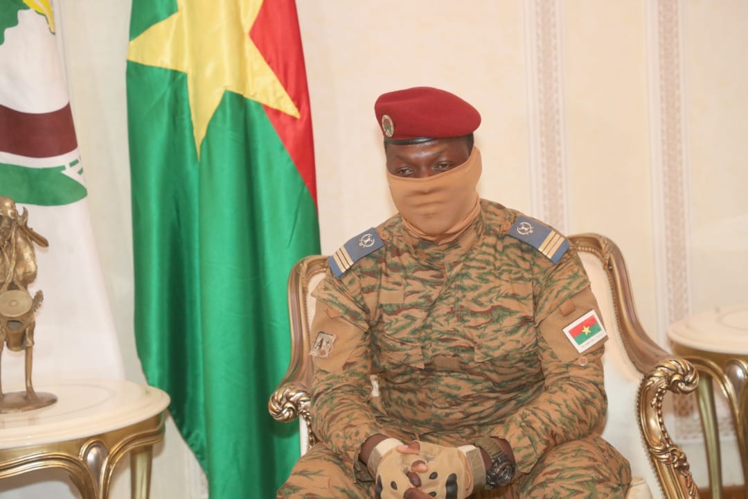 Burkina Faso army captain Ibrahim Traoré is seated in front of the Burkina Faso flag. His mouth and nose are covered with a gaiter.
